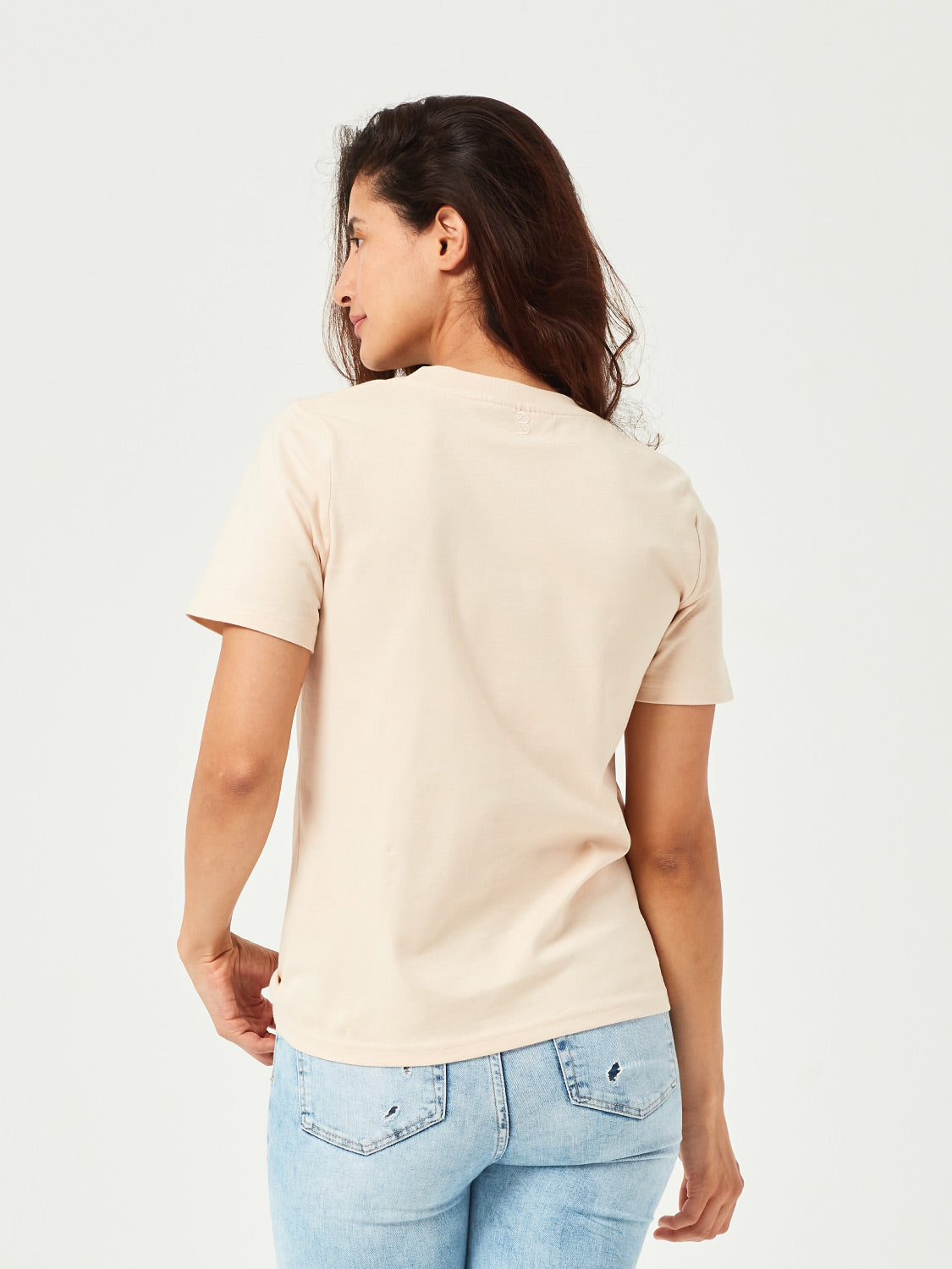 Frau in sand farbenen T-Shirt und hellblauen zerrissenen Jeans, steht mit dem Rücken zur Kamera, weinrote Sneakers, vor weißem Hintergrund.