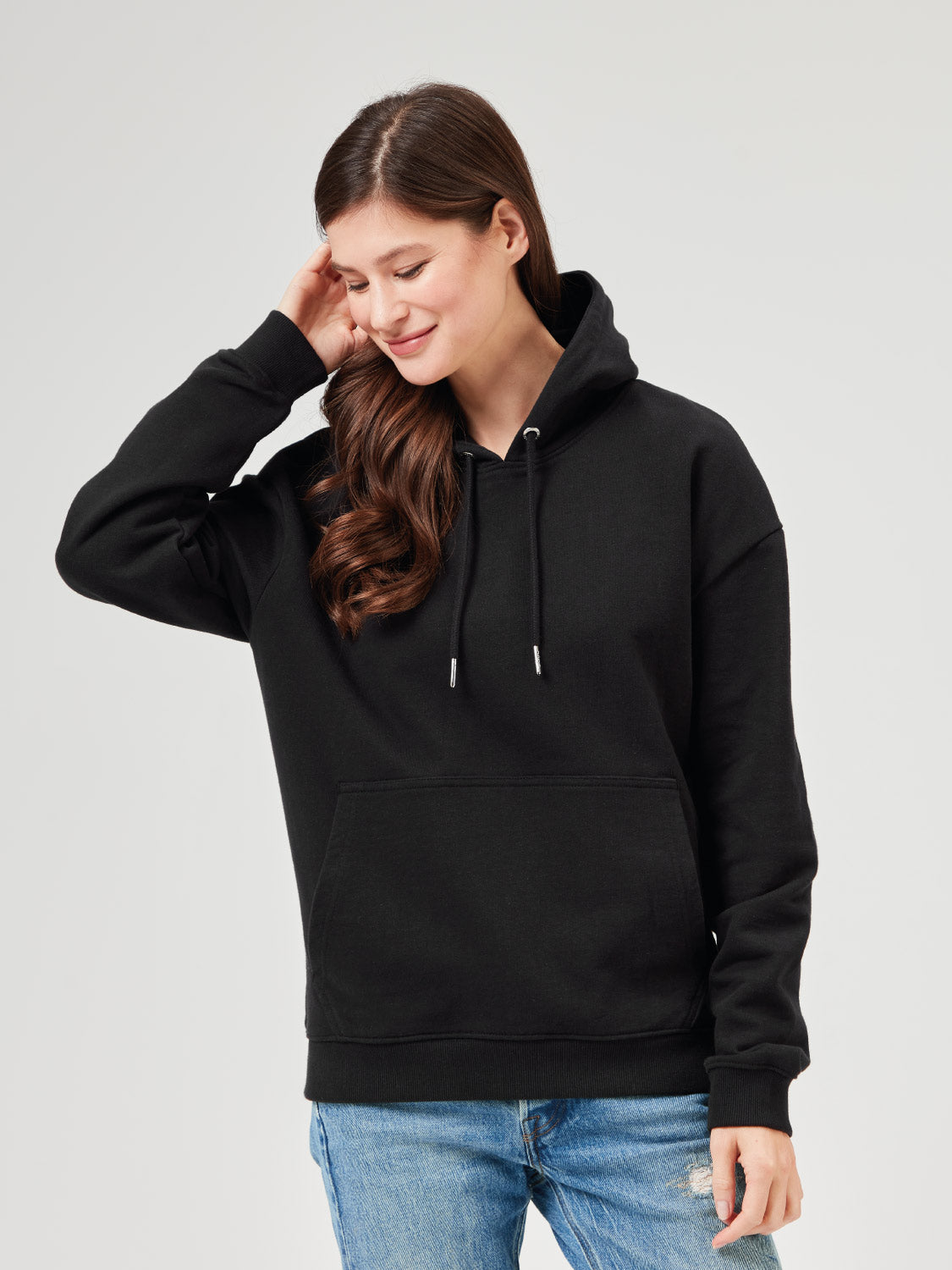 Frau trägt schwarzen Unisex Hoodie aus Bio-Baumwolle mit Kapuze und Kängurutasche. Kombiniert mit hellblauer Jeans und schwarzen Boots für lässige und stylische Casual Outfits. Nachhaltige Mode für Frauen.