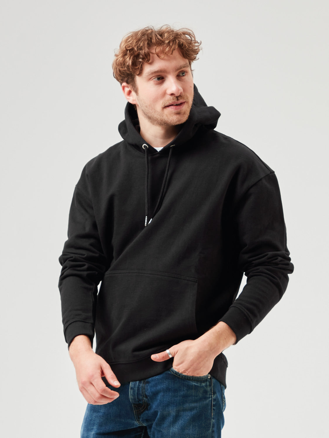 Mann trägt schwarzen Unisex Hoodie aus Bio-Baumwolle mit Kapuze und Kängurutasche, kombiniert mit hellblauer Jeans. Bequemer, nachhaltiger Hoodie für Freizeitmode, Streetwear und moderne Casual Styles.