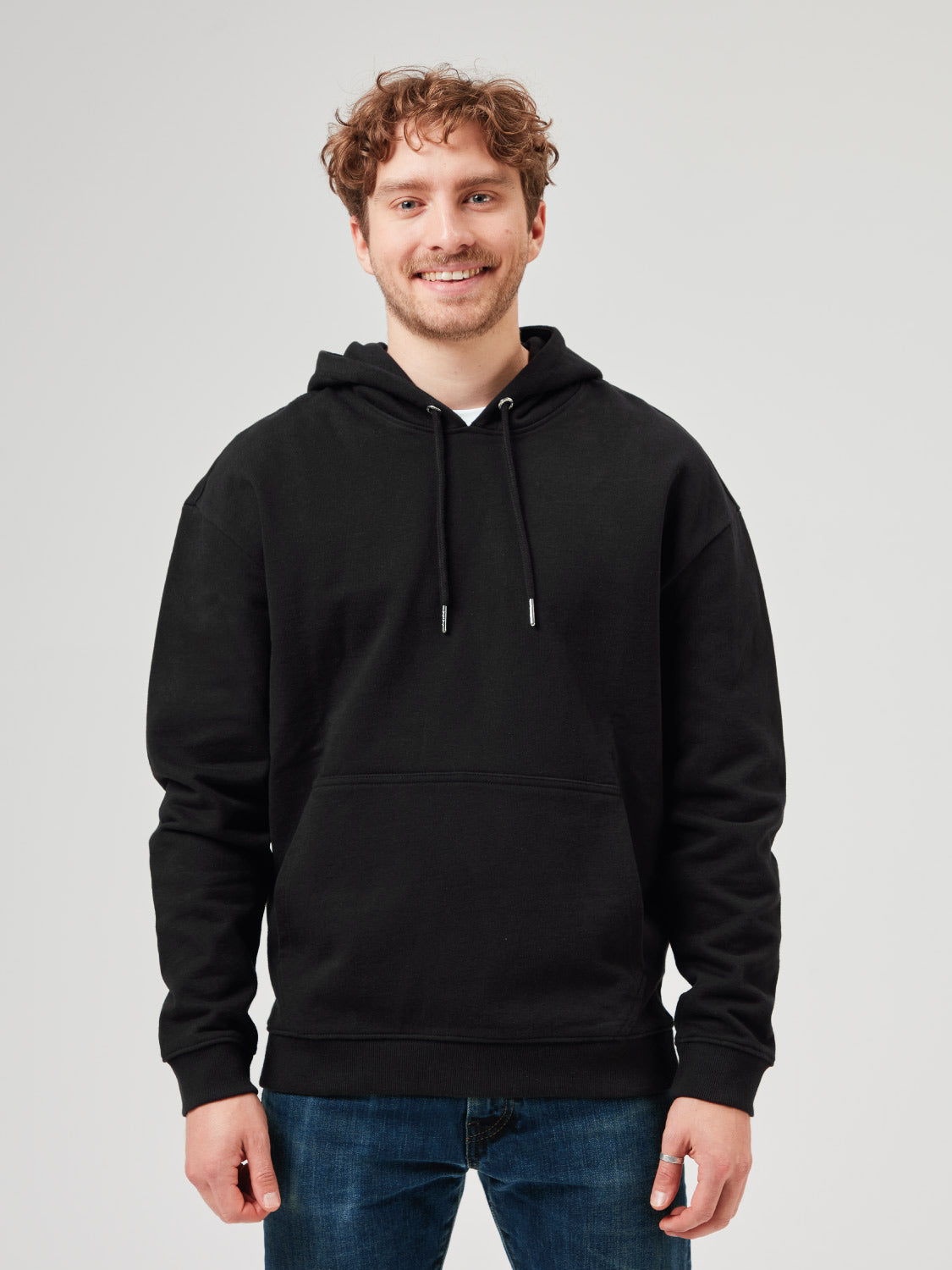 Mann trägt schwarzen Unisex Hoodie aus Bio-Baumwolle mit Kapuze und Kängurutasche, kombiniert mit hellblauer Jeans. Bequemer, nachhaltiger Hoodie für Freizeitmode, Streetwear und moderne Casual Styles.
