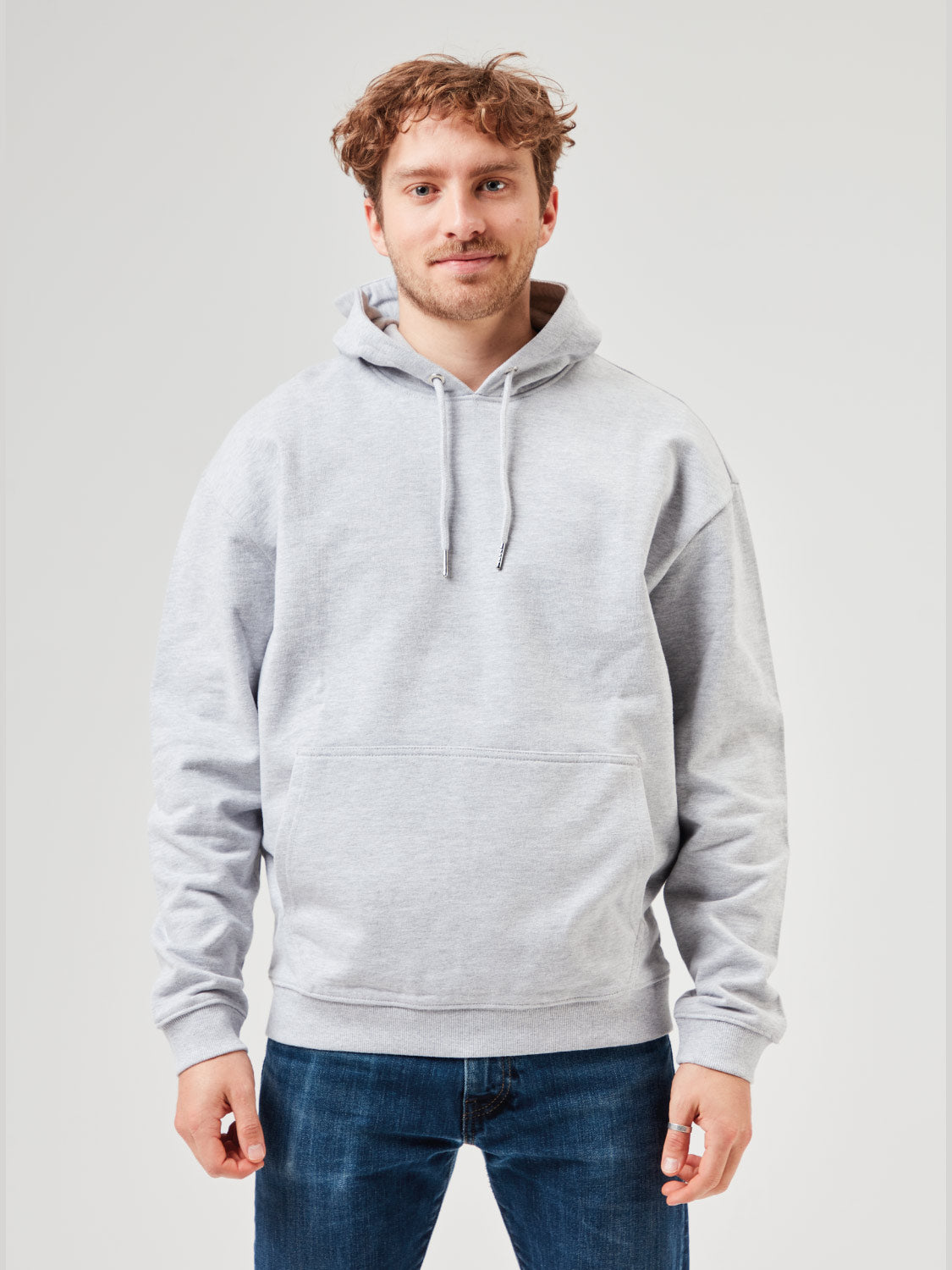 Mann trägt grauen Unisex Hoodie aus Bio-Baumwolle mit Kapuze und Kängurutasche. Kombiniert mit blauen Jeans für lässige Freizeitlooks und moderne Streetwear. Nachhaltige Mode für Männer.