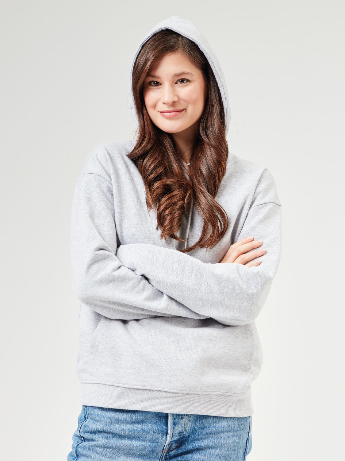 Frau trägt grau melangen Unisex Hoodie aus Bio-Baumwolle mit Kapuze und Kängurutasche. Stylische Kombination mit blauen Jeans für Casual Outfits und umweltfreundliche Streetwear.