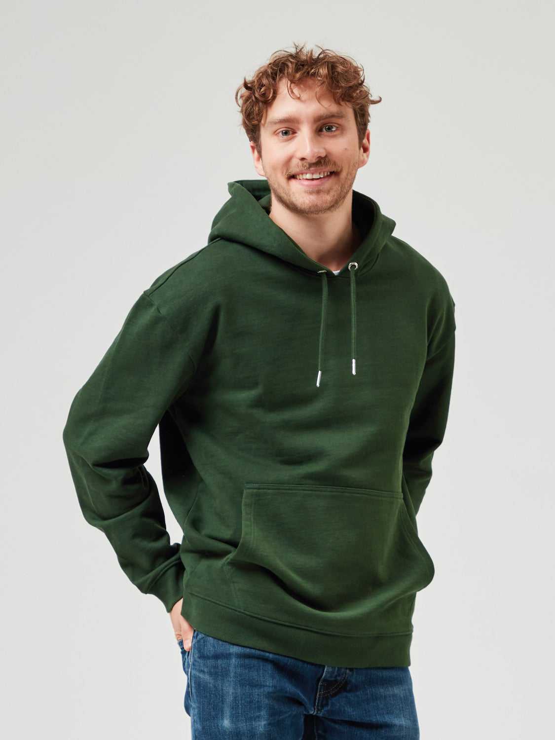 Unisex Hoodie in Dunkelgrün aus Bio-Baumwolle mit Kapuze und Kängurutasche. Lässiger Kapuzenpullover kombiniert mit blauer Jeans. Nachhaltige Mode für Männer und Frauen, perfekt für Freizeitlooks und Streetwear.